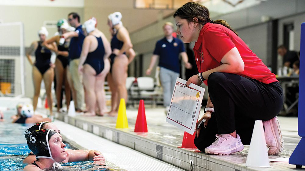 water polo coach kneeling at edge of the pool talking to females in pool